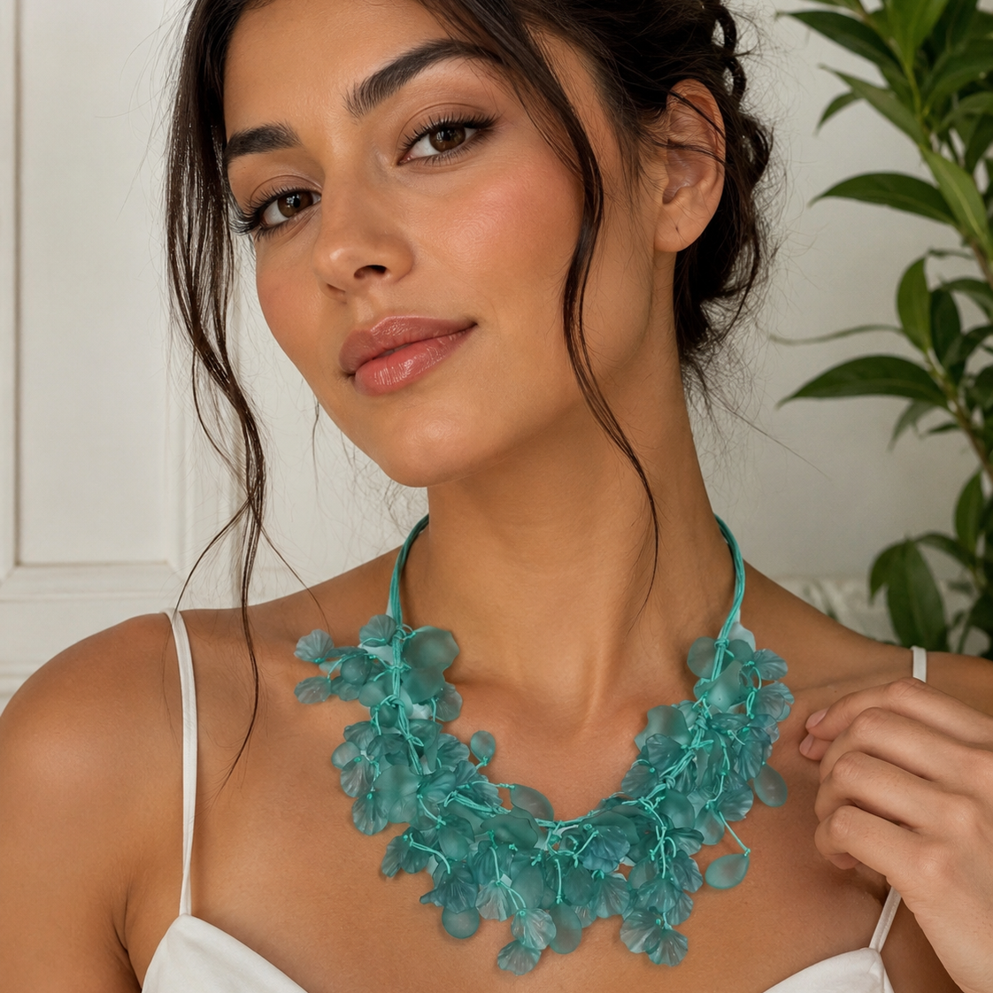 A woman wearing a chunky, layered turquoise floral statement necklace over a white spaghetti-strap top, with a soft-focus indoor background featuring a white wall and green leaves.