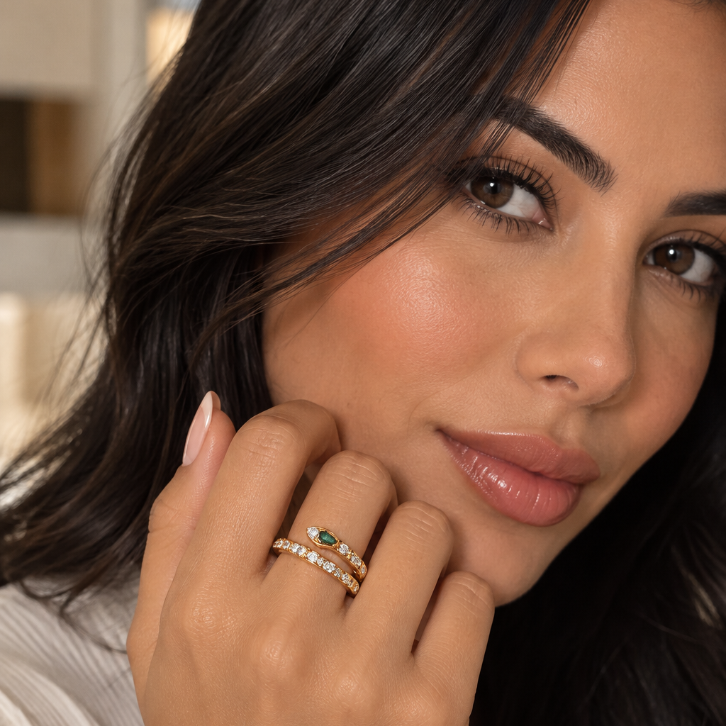 A close-up lifestyle shot of a woman wearing a double stack ring set. The set features a central oval green stone surrounded by small clear stones on a gold band, worn over a matching gold band with tiny clear stones.