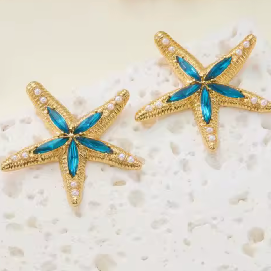 A pair of gold starfish-shaped earrings with blue marquise-cut center stones and small white pearl-like accents, displayed on a textured white surface against a warm cream background.