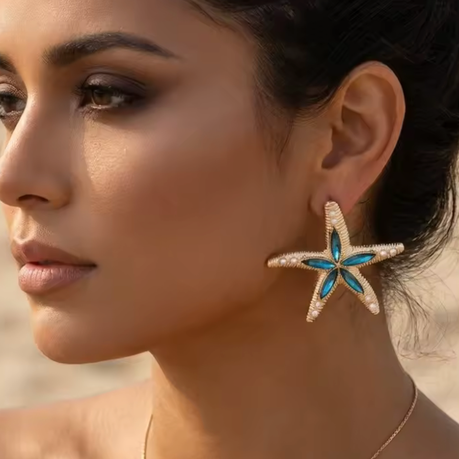 A close-up of a woman wearing a gold starfish-shaped earring with a blue and turquoise center, set against a softly blurred warm background.