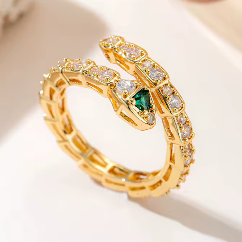 A gold snake ring with a green emerald-cut center stone and clear accent stones, photographed on a clean light background.