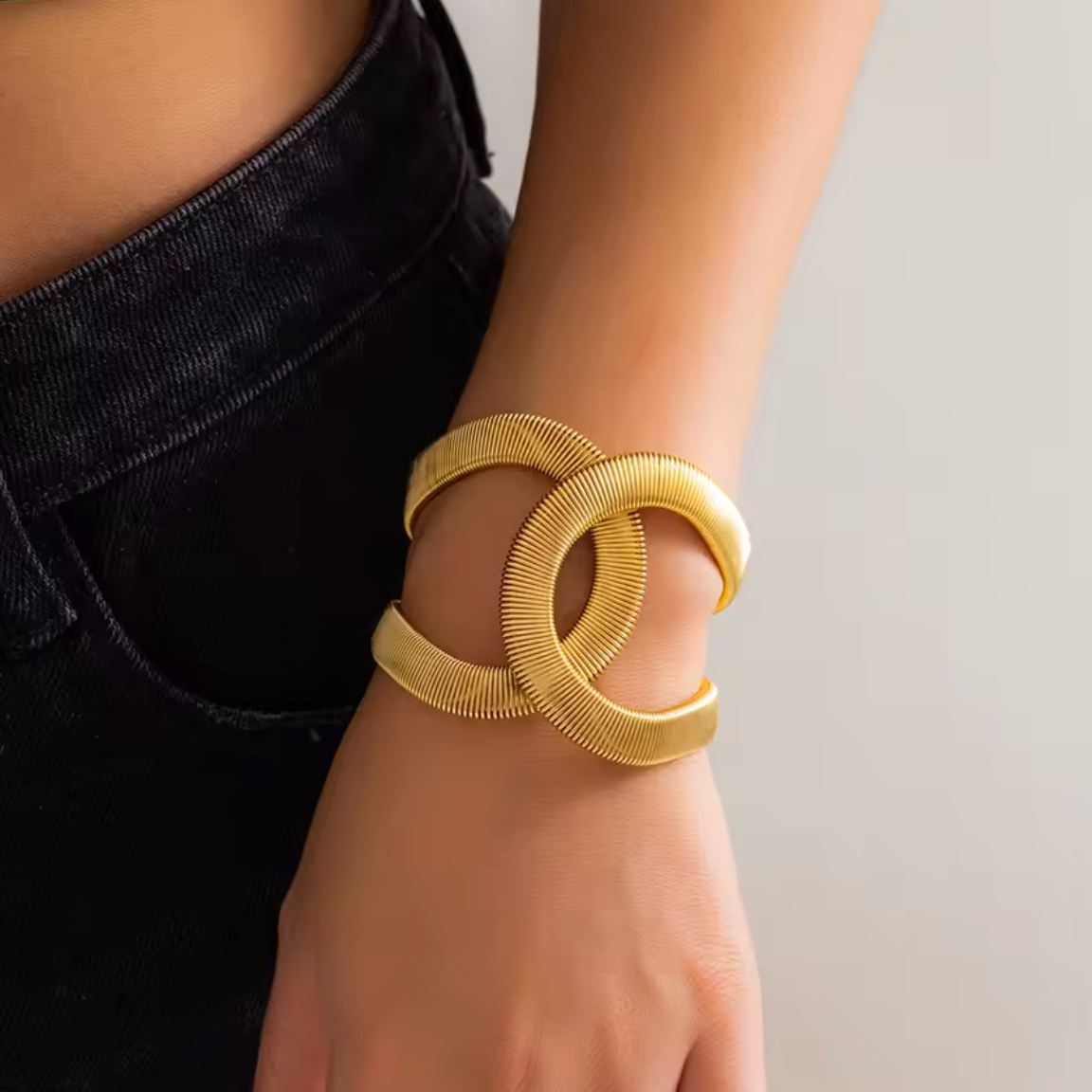 A close-up of a person's wrist wearing a chunky, double-loop interlocking cuff bracelet in polished yellow gold. The person is wearing black denim jeans, and the background is a clean, neutral light gray.