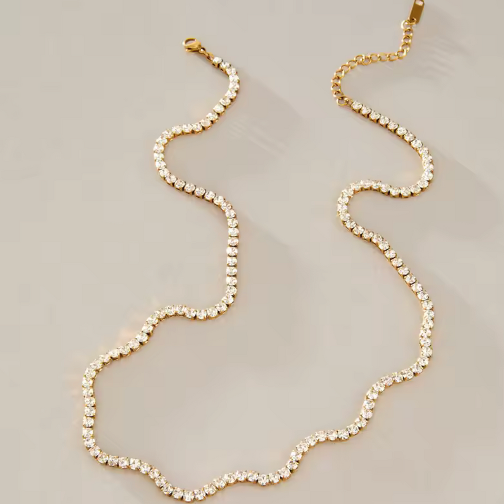 A gold chain necklace with a wavy, scalloped edge and clear rhinestone-like embellishments, displayed on a neutral light gray background.