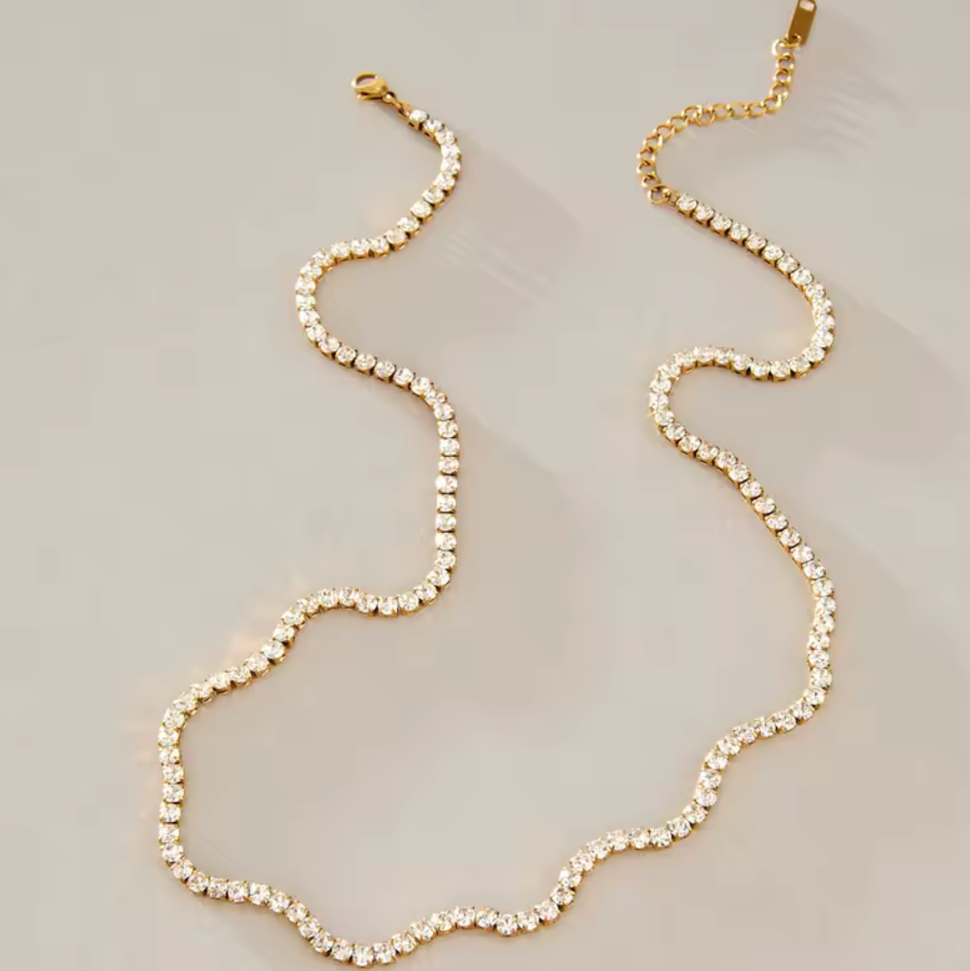 A gold chain necklace with a wavy, scalloped edge and clear rhinestone-like embellishments, displayed on a neutral light gray background.