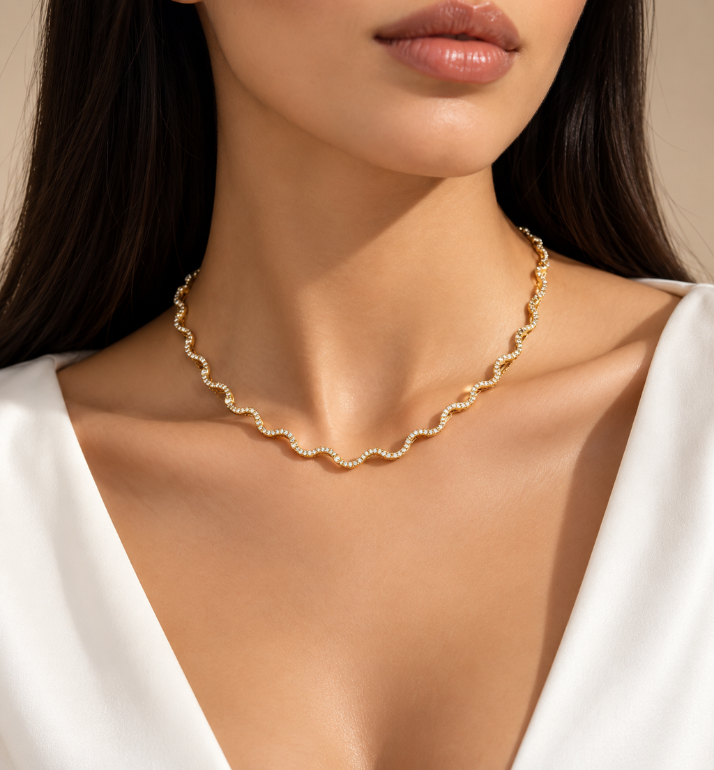 A close-up of a model wearing a delicate gold wavy chain necklace with small clear stones, paired with a white V-neck top against a warm beige background.